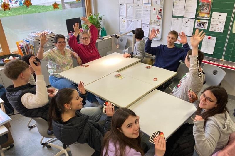 Mehrere Schüler und eine Erwachsene Person sitzen um zusammengestellte Tische, heben die Hände und halten Spielkarten, während sie in einem hellen Klassenraum spielen.