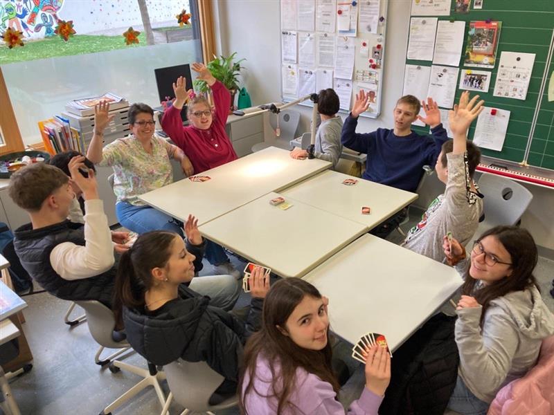 Mehrere Schüler und eine Erwachsene Person sitzen um zusammengestellte Tische, heben die Hände und halten Spielkarten, während sie in einem hellen Klassenraum spielen.