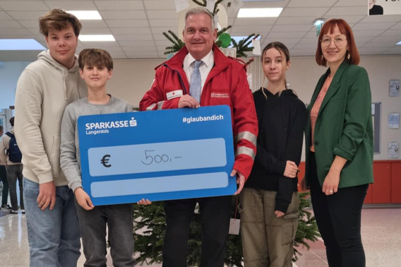 Lehrer und Lehrerin mit drei Schüler*innen und € 500 Spendentafel, direkt in die Kamera blickend.