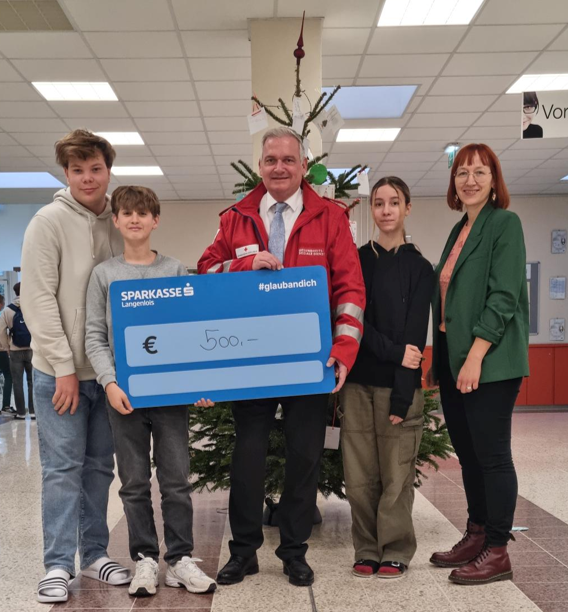 Lehrer und Lehrerin mit drei Schüler*innen und € 500 Spendentafel, direkt in die Kamera blickend.
