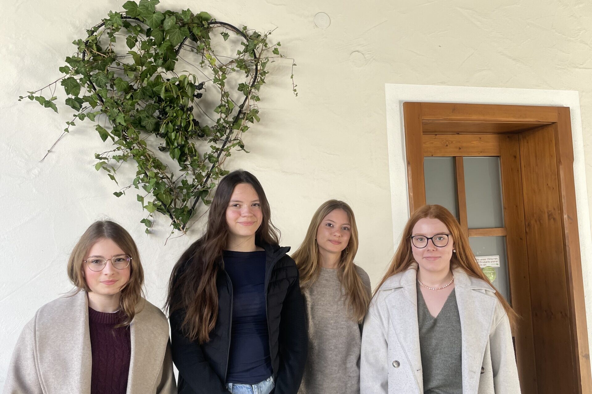 Vier Schülerinnen stehen vor einer Wand mit herzförmiger Pflanzen-Deko neben einer Holztür
