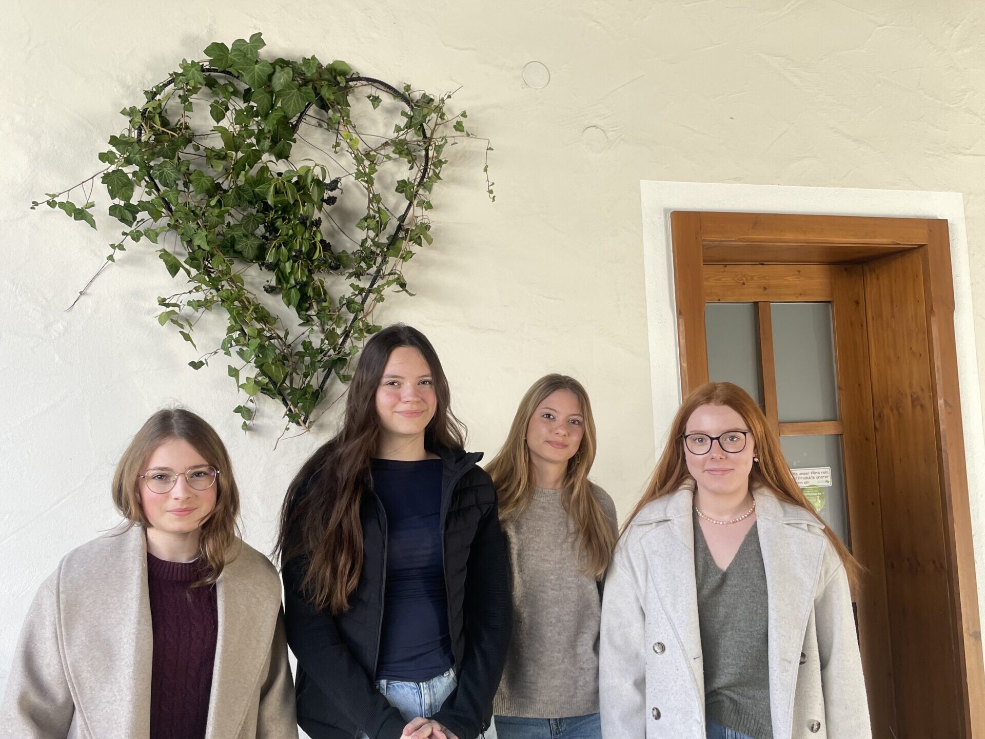 Vier Schülerinnen stehen vor einer Wand mit herzförmiger Pflanzen-Deko neben einer Holztür