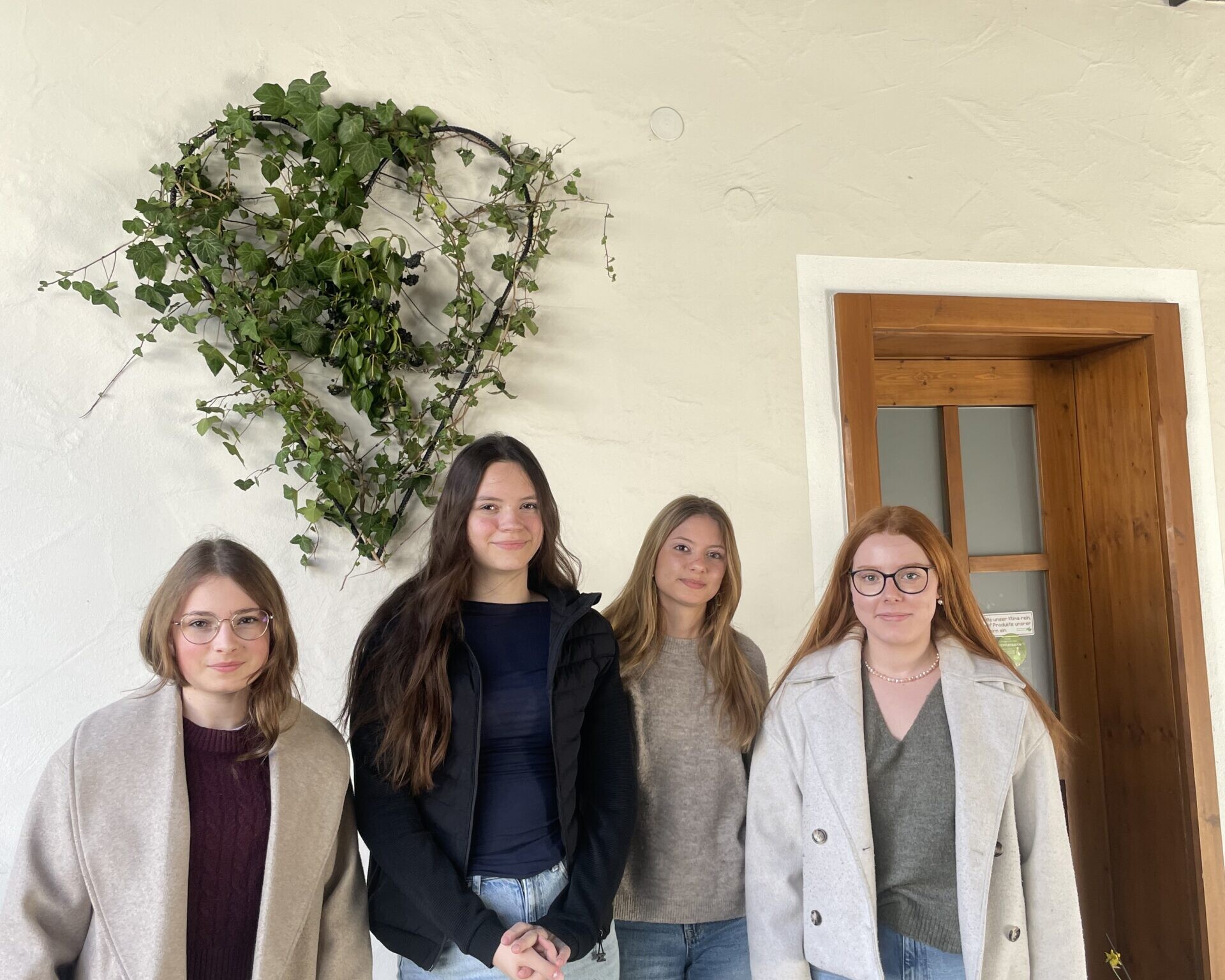 Vier Schülerinnen stehen vor einer Wand mit herzförmiger Pflanzen-Deko neben einer Holztür