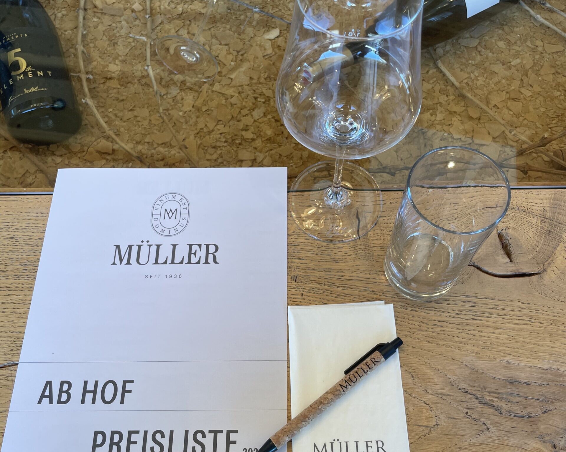 Weinglas, Wasserglas und Preisliste auf Holztisch bei einer Weinverkostung, mit Flaschen und Reben im Hintergrund