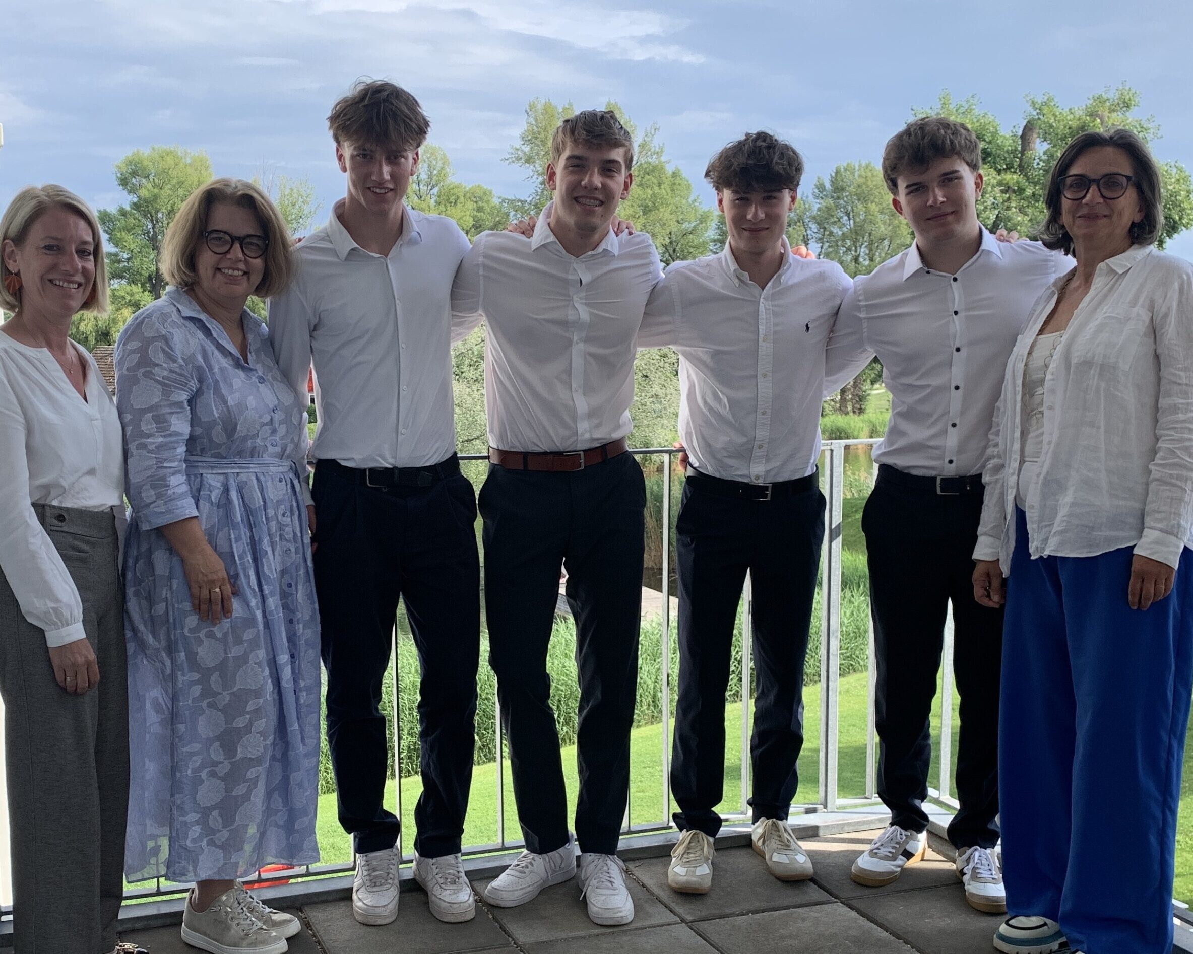 Vier Schüler und drei Lehrkräfte stehen eng beieinander auf einem Balkon und lächeln in die Kamera. Im Hintergrund sind Bäume, Wiesen und ein bewölkter Himmel zu sehen.