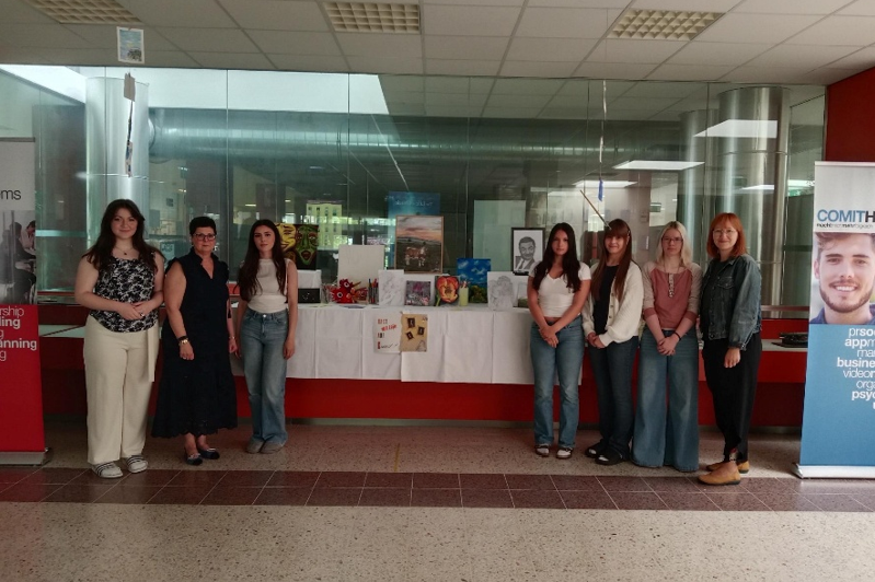 Fünf Schülerinnen präsentieren gemeinsam mit einer Lehrerin und einer Direktorin eine Ausstellung mit Kunstwerken und Projekten in einem Schulgebäude.