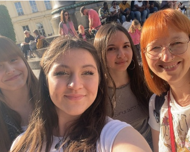 Drei Schülerinnen und eine Lehrerin lächeln für ein Selfie auf einem sonnigen Platz im Freien.
