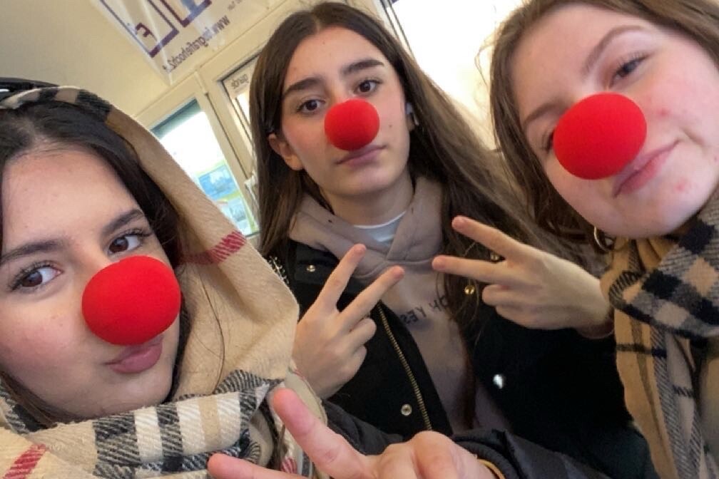 3 Personen mit roten Nasen auf ihrer Nase, sehen direkt in die Kamera und lächeln dabei, eine Frau hält dabei eine Spendendose