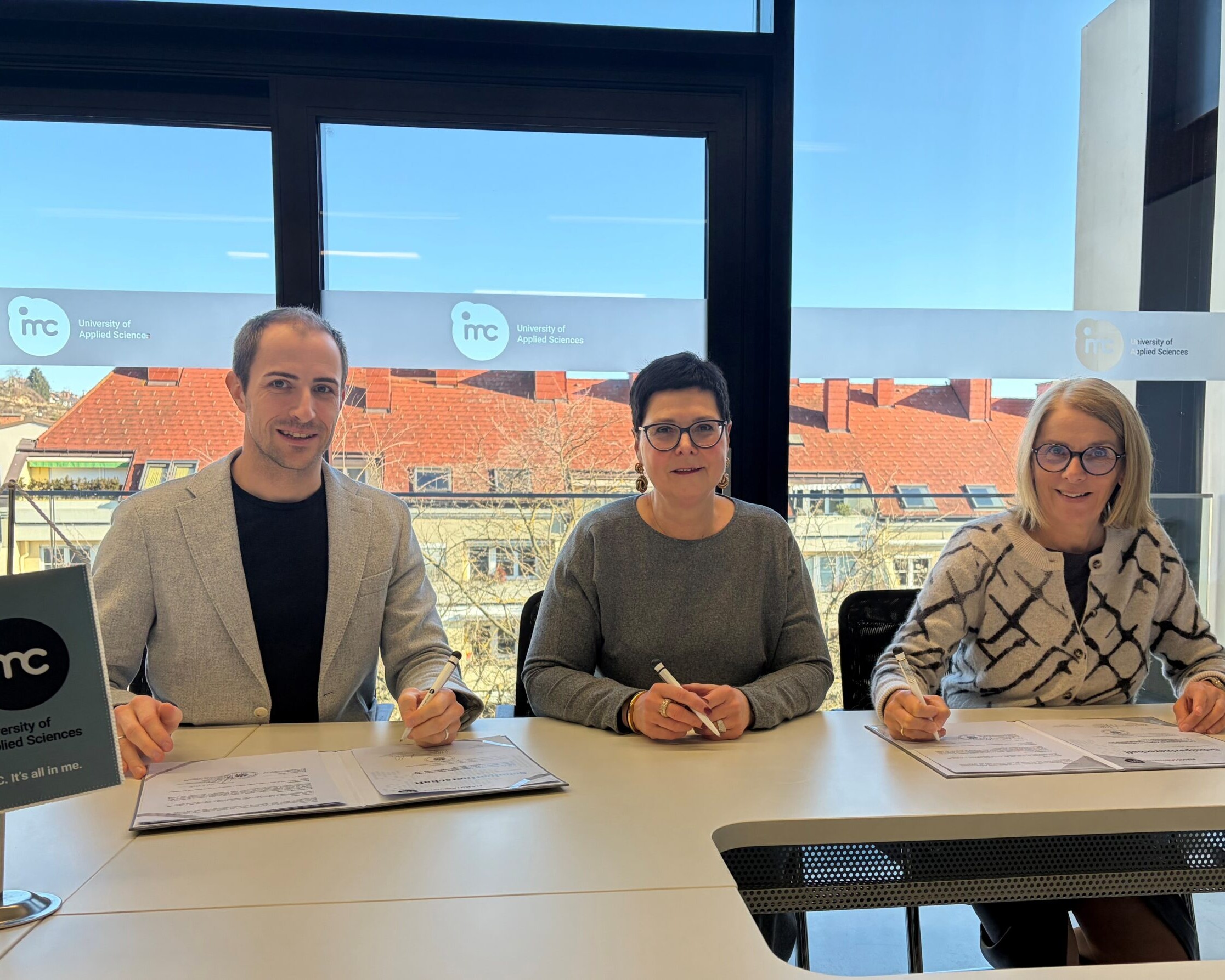 Drei Personen sitzen an einem Tisch und unterzeichnen Dokumente in einem Besprechungsraum der IMC University of Applied Sciences.