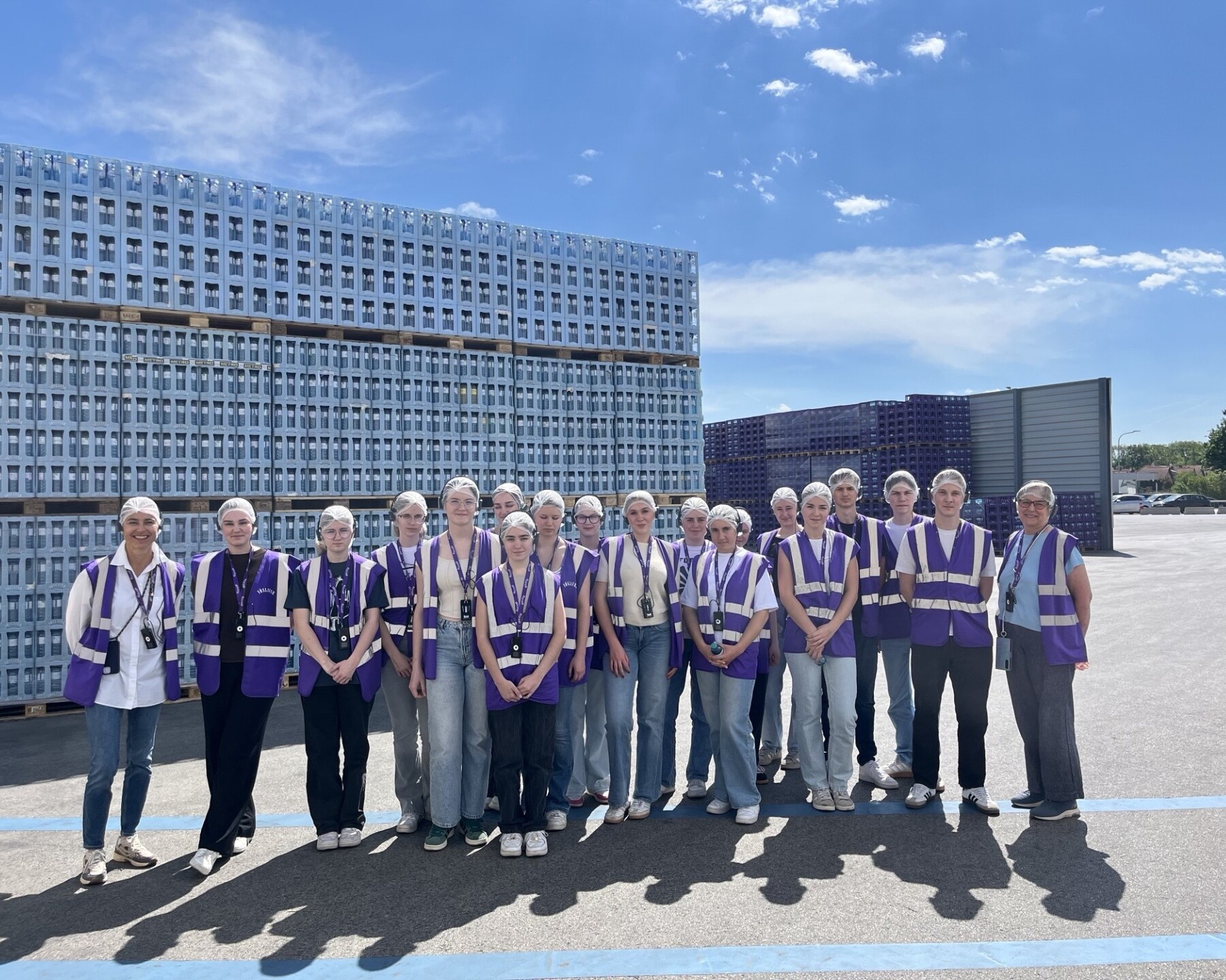 Eine Gruppe von Personen mit Schutzhauben und violetten Warnwesten steht vor gestapelten Kisten auf einem Industriegelände unter blauem Himmel.