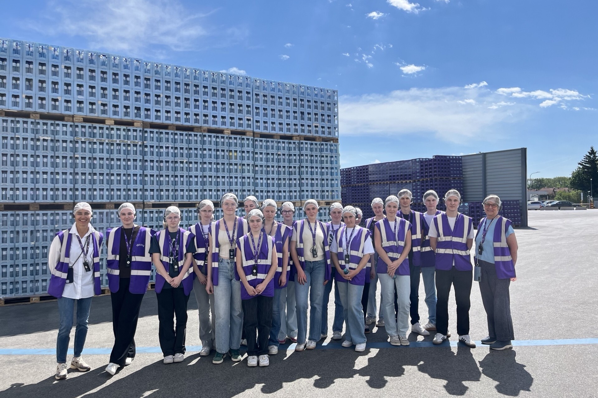 Eine Gruppe von Personen mit Schutzhauben und violetten Warnwesten steht vor gestapelten Kisten auf einem Industriegelände unter blauem Himmel.