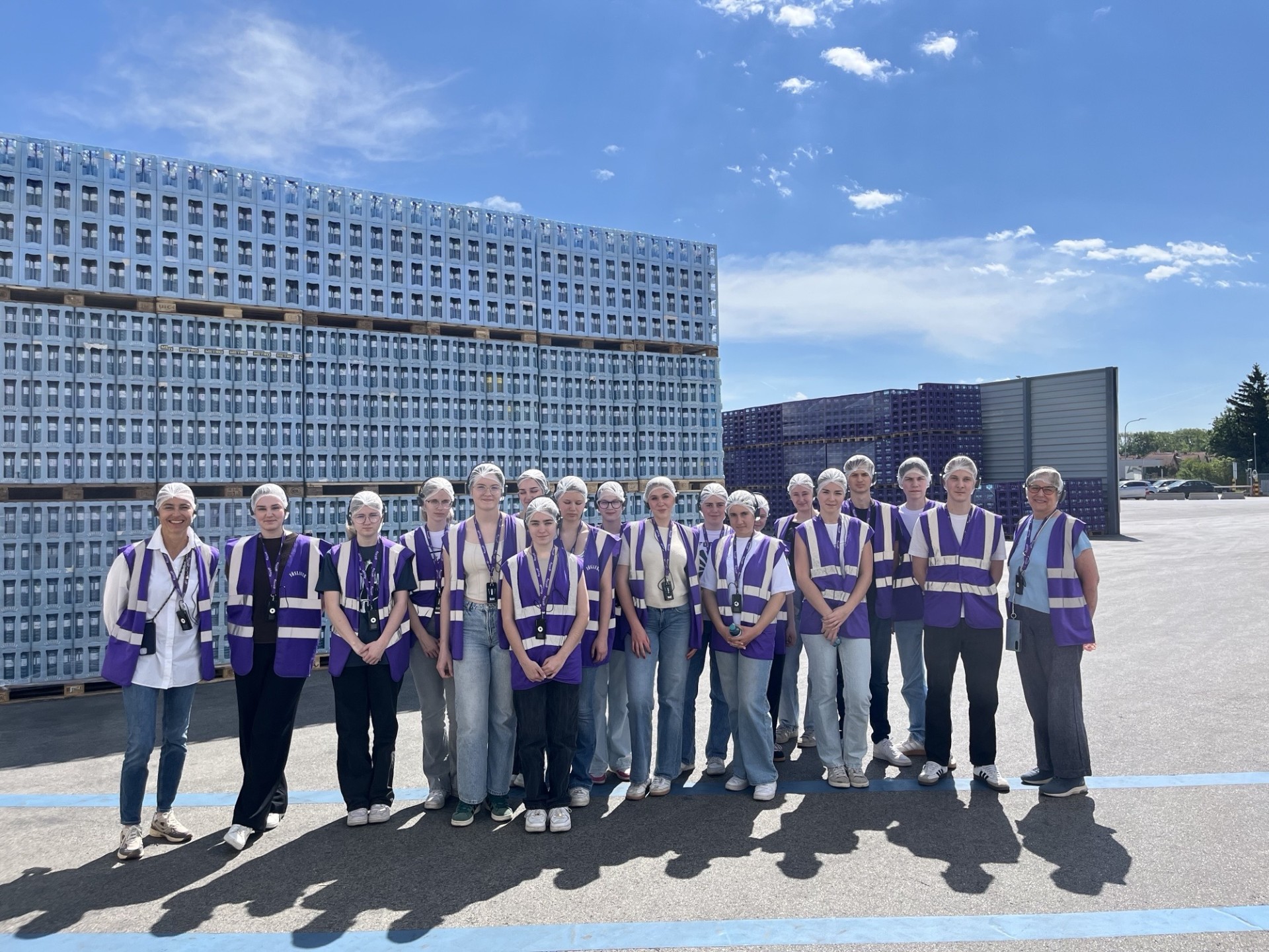 Eine Gruppe von Personen mit Schutzhauben und violetten Warnwesten steht vor gestapelten Kisten auf einem Industriegelände unter blauem Himmel.