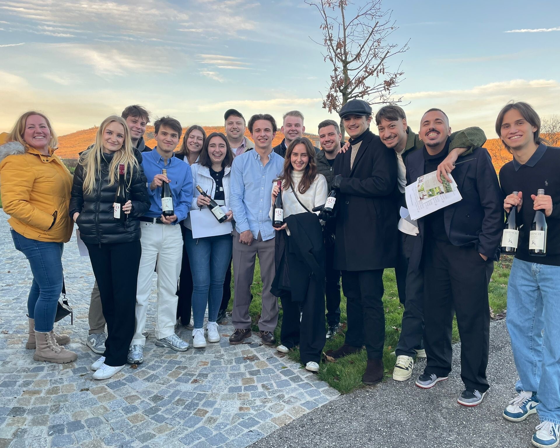 15 Personen mit Weinflaschen in der Hand, draußen stehend und direkt in die Kamera schauend.