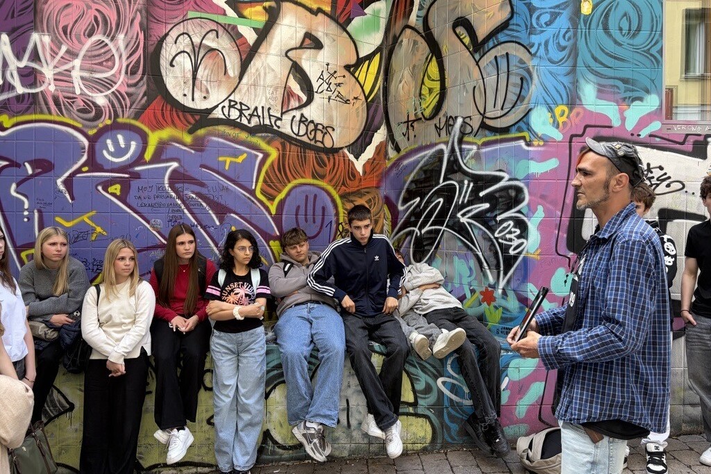 Eine Gruppe Jugendlicher sitzt vor einer bunten Graffitiwand und hört einem Mann bei einer Stadtführung zu.