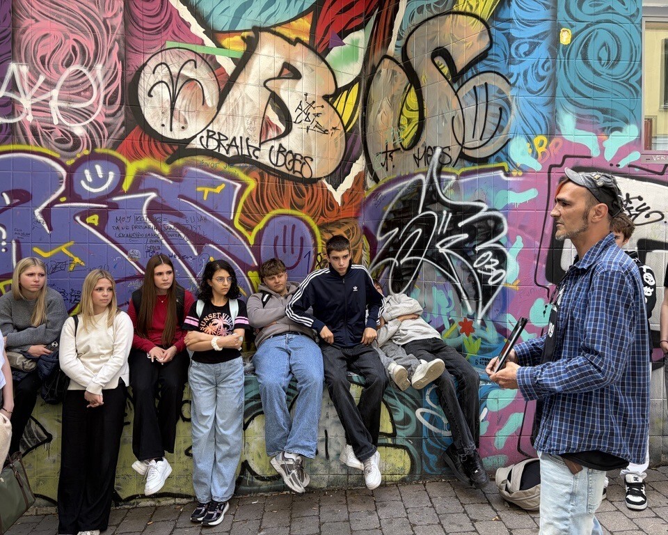 Eine Gruppe Jugendlicher sitzt vor einer bunten Graffitiwand und hört einem Mann bei einer Stadtführung zu.