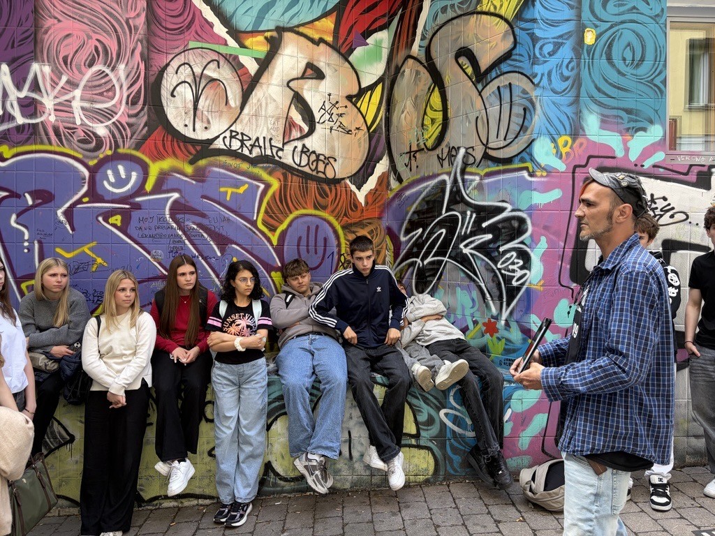 Eine Gruppe Jugendlicher sitzt vor einer bunten Graffitiwand und hört einem Mann bei einer Stadtführung zu.
