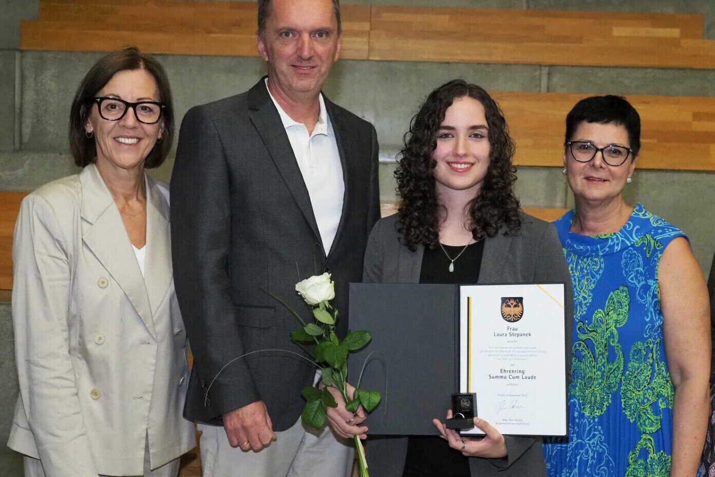 Vier Personen stehen nebeneinander, eine junge Frau steht mit offener Urkunde und weißer Rose in ihren Händen, die anderen 3 Personen stehen links und rechts von ihr und alle schauen in die Kamera und lächeln