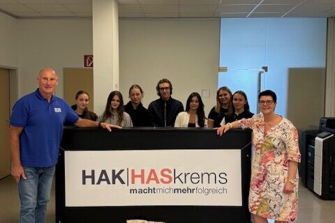 7 Schüler*innen mit Direktorin und 1 Mitarbeiter von simply strong stehend vor einem Pult wo man das HAK|HASkrems Logo sieht, die Personen sehen direkt in die Kamera und lächeln