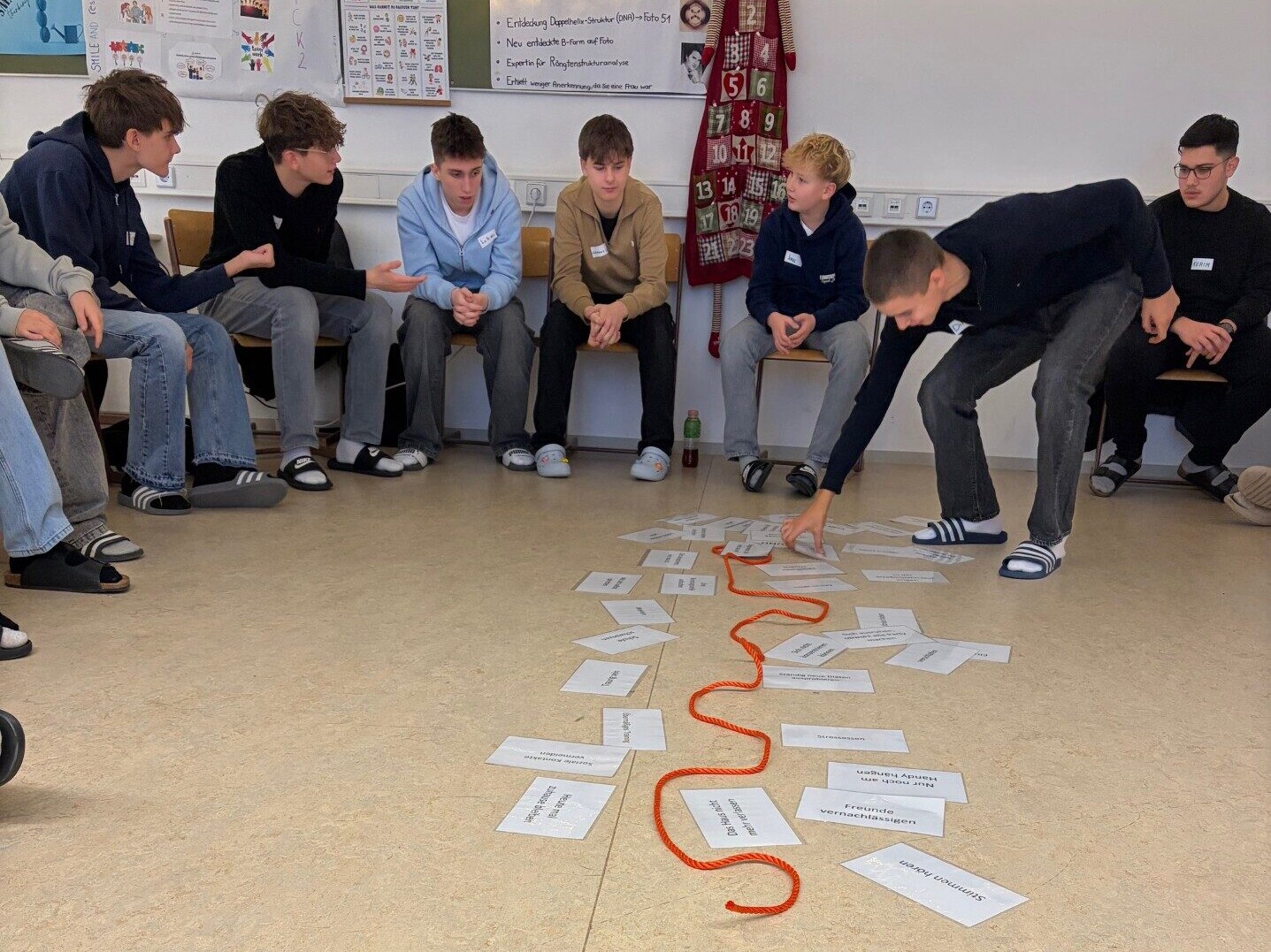 Sechs sitzende Sch&uuml;ler in einem Halbkreis, w&auml;hrend ein Sch&uuml;ler einen Zettel in die Mitte des Kreises zu anderen Zetteln und einer Schnur hinlegt.