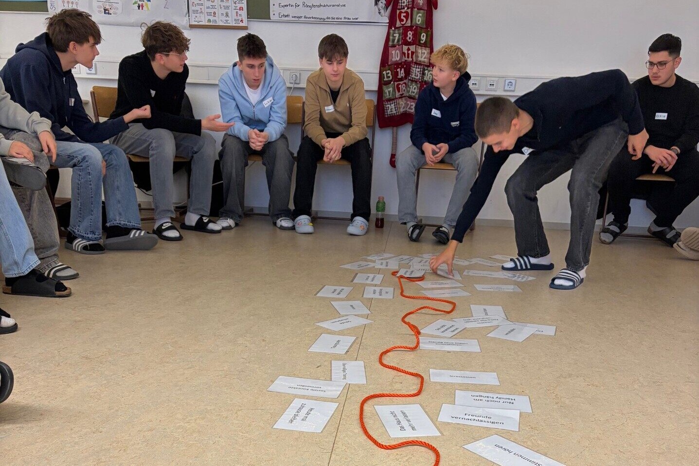 Sechs sitzende Schüler in einem Halbkreis, während ein Schüler einen Zettel in die Mitte des Kreises zu anderen Zetteln und einer Schnur hinlegt.