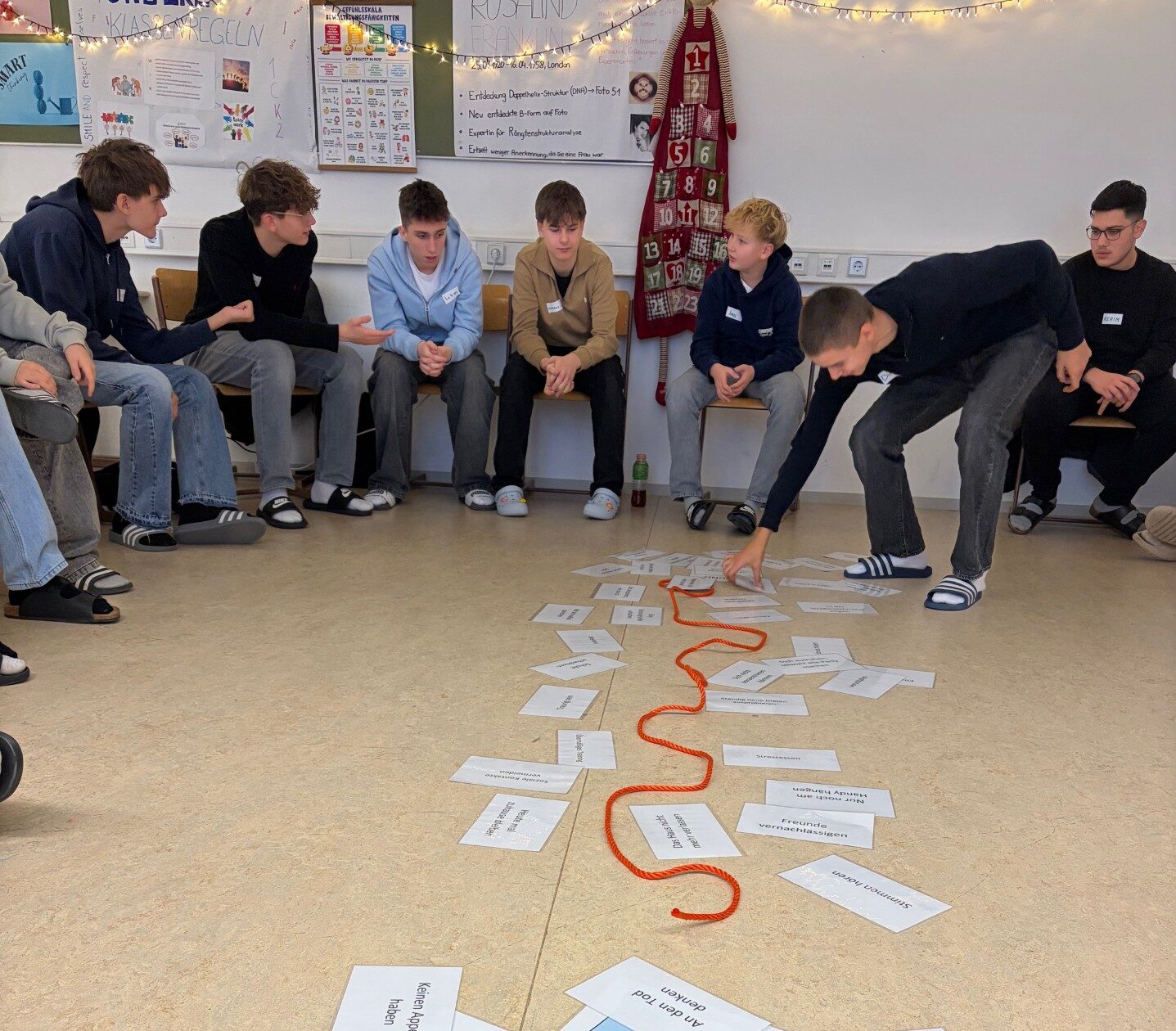 Sechs sitzende Schüler in einem Halbkreis, während ein Schüler einen Zettel in die Mitte des Kreises zu anderen Zetteln und einer Schnur hinlegt.